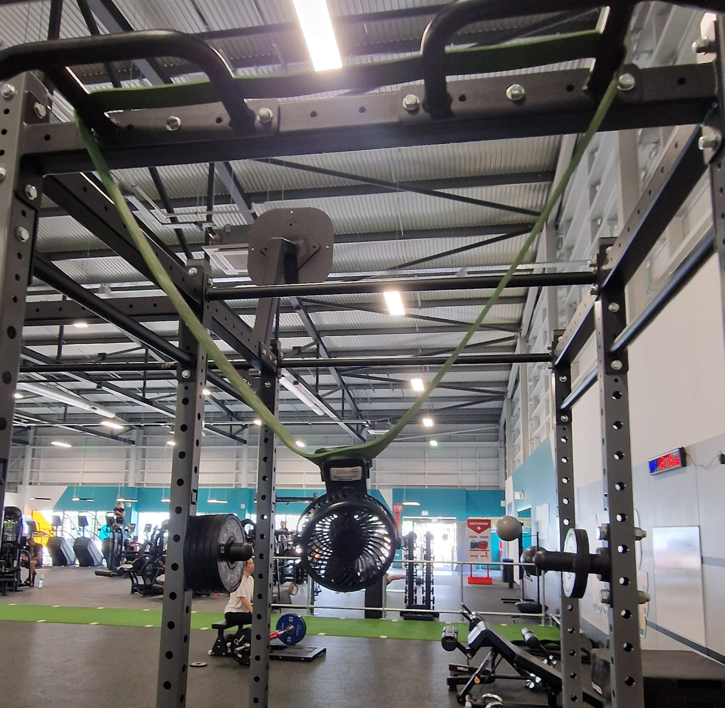 A green resistance band hangs from the pull up bars above a squat rack in a gym, with a portable USB fan clipped onto it blowing air towards the camera.