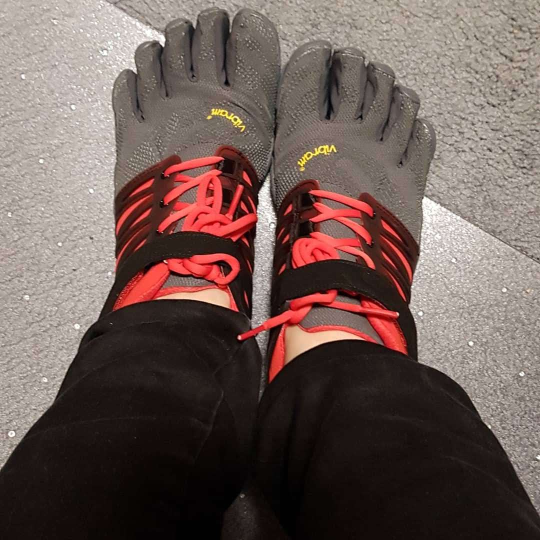 Photo of two feet wearing black, grey and red Vibram five fingers Train shoes which have individual toes and lace up with a velcro strap over the top.